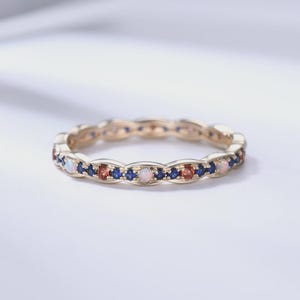 Könnte beinhalten: Ein goldener Ring mit einem sich wiederholenden Muster aus Edelsteinen. Der Ring hat abwechselnd blaue, orangefarbene und weiße Edelsteine, die in ein goldenes Band gefasst sind. Der Ring ist ein zartes Schmuckstück.