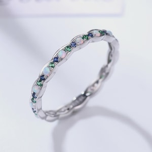 Pode incluir: Um anel de prata com um design ondulado, cravejado com pedras preciosas de opala, verdes e azuis alternadas. O anel é uma joia delicada, perfeita para uso diário ou ocasiões especiais.