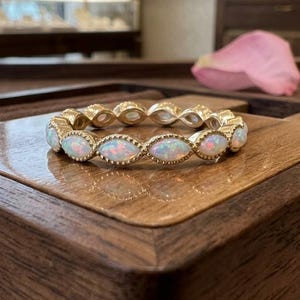 May include: A gold ring with a series of oval-shaped opals set in a repeating pattern. The opals have a milky white base with flashes of color. The ring is displayed on a wooden surface.