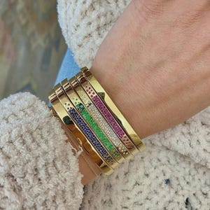 May include: A collection of gold bangles, some adorned with colorful gemstones. The bangles are stacked on a wrist, showcasing a variety of designs. The gemstones include blue, green, white, and pink, adding a vibrant touch to the gold. The bangles are approximately 0.25 inches wide.