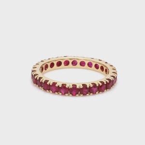 Peut inclure: Une bague d'éternité dorée ornée de nombreux rubis rouges vifs. La monture de la bague est un cercle continu de pierres rouges, créant un look audacieux et élégant. La bague est présentée sur un fond clair et uni.