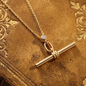 Puede incluir: Un collar de oro con una cadena fina conectada a un pequeño engaste redondo con una piedra transparente. Debajo de la piedra, un eslabón ovalado de oro se une a un colgante de barra horizontal de oro, creando un diseño de cierre de barra.