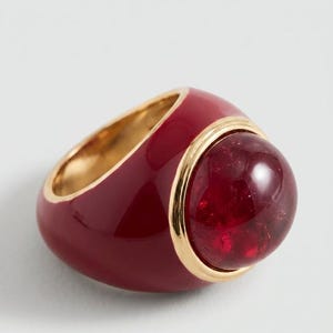 May include: A bold ring featuring a deep red, smooth enamel band with a polished gold-tone metal interior and trim. The centre is set with a large, translucent, dark red cabochon stone that is framed by a thin, circular gold-tone metal border.