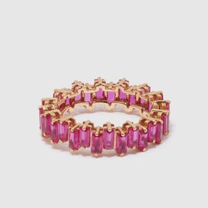 May include: A gold-colored ring featuring a continuous band of rectangular, vibrant pink gemstones. The gemstones are set closely together, creating a full, eye-catching design. The ring is displayed against a plain white background.