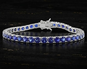 May include: A silver-toned tennis bracelet featuring a continuous row of round, deep blue stones set in a metallic frame. The bracelet includes a secure, hinged box clasp mechanism at the center, connecting the two ends of the circular chain.