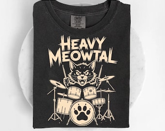 May include: Dark gray t-shirt with a cream-colored graphic. The graphic features a black cat playing a drum set with the words "HEAVY MEOWTAL" above it. The bass drum has a paw print design.