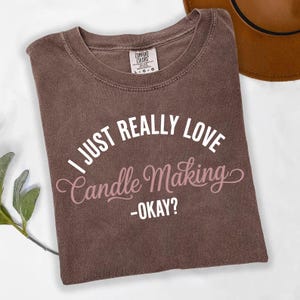 Könnte beinhalten: Ein braunes T-Shirt mit dem Text "I JUST REALLY LOVE Candle Making - OKAY?" in weißer und rosa Schrift. Das Shirt hat einen Rundhalsausschnitt und ein Comfort Colors-Etikett.