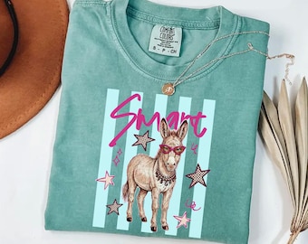 May include: A sage green short-sleeve t-shirt featuring a graphic of a donkey wearing pink glasses and a beaded necklace. The design includes the word Smart in pink script, light blue vertical stripes, and various star shapes.