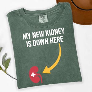 May include: A sage green t-shirt with the text "MY NEW KIDNEY IS DOWN HERE" in white. A red kidney graphic with a white cross is positioned below the text, with a yellow arrow pointing towards it.