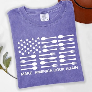 Puede incluir: Una camiseta morada Comfort Colors con un diseño gráfico blanco de la bandera estadounidense hecho con cucharas, tenedores y cuchillos. El texto "MAKE AMERICA COOK AGAIN" está impreso debajo. La camiseta está doblada y tiene una etiqueta.
