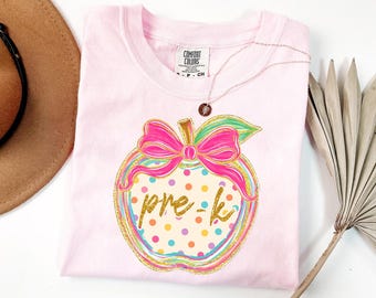 May include: A light pink t-shirt featuring a graphic of an apple with a bright pink bow. The apple has a cream-coloured background with multi-coloured polka dots and the text pre-k written in gold glitter script across the centre.