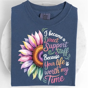 May include: A navy blue t-shirt with a vibrant sunflower graphic. The design includes a half-sunflower with pink, yellow, and green petals. Text on the shirt says: "I became a Direct Support Staff Because Your life is worth my Time."