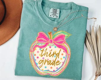 May include: A sage green t-shirt featuring a graphic of an apple with a bright pink bow. The apple has colourful polka dots and the gold glitter text third grade written across the centre. The shirt has a Comfort Colors brand label at the neck.