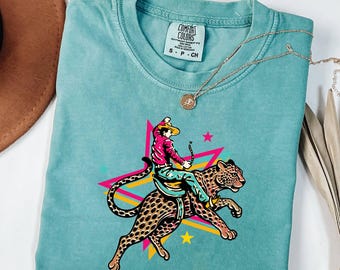May include: A teal crewneck t-shirt featuring a graphic of a person in a cowboy hat and pink shirt riding a leopard. The design includes a multi-coloured star background with pink, yellow, and blue lines. The shirt has a Comfort Colors brand label.