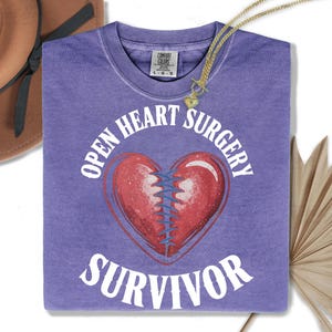 May include: A purple t-shirt with the words "OPEN HEART SURGERY SURVIVOR" in white. A red heart with blue stitching is in the center. A gold necklace with a heart pendant is draped over the shirt. A brown hat is in the upper left corner.