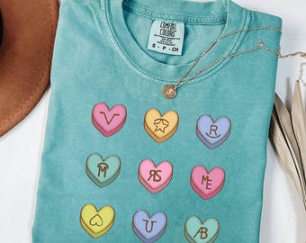 May include: A teal short-sleeve t-shirt featuring a graphic print of nine colourful candy hearts arranged in a grid. Each heart displays a unique letter or symbol, including a star, a heart outline, and various letter combinations like V, R, M, RS, ME, U, and AB.
