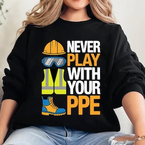 Op de afbeelding: Zwart sweatshirt met de tekst "NEVER PLAY WITH YOUR PPE" in wit en oranje. Het ontwerp bevat illustraties van een gele helm, een veiligheidsbril, een reflecterend vest en werkschoenen.
