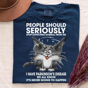 May include: A navy blue t-shirt with the text "PEOPLE SHOULD SERIOUSLY STOP EXPECTING NORMAL FROM ME" in white. Below the text is a cartoon cat with its tongue out. The text "I HAVE PARKINSON'S DISEASE WE ALL KNOW IT'S NEVER GOING TO HAPPEN" is below the cat.