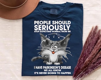 May include: A navy blue t-shirt featuring the text "PEOPLE SHOULD SERIOUSLY STOP EXPECTING NORMAL FROM ME" in white. A cartoon cat with its tongue out is below the text. The text "I HAVE PARKINSON'S DISEASE WE ALL KNOW IT'S NEVER GOING TO HAPPEN" is below the cat.