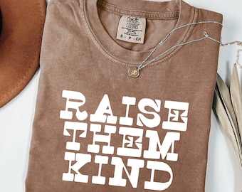 May include: A brown short-sleeved t-shirt with a crew neckline and a Comfort Colors brand label at the inner collar. The front features the white text RAISE THEM KIND in a bold, blocky font. A silver-toned necklace with a small circular pendant.