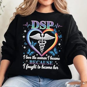 May include: Black sweatshirt with a colourful heart-shaped design featuring butterflies, a medical symbol, and the letters "DSP." The text "I love the woman I became BECAUSE I fought to become her" is also displayed.