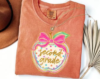 May include: A terracotta-coloured short-sleeve t-shirt featuring a graphic of an apple with a bright pink bow. The apple has a cream-coloured background with multi-coloured polka dots and the gold glitter text second grade.