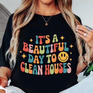 May include: Black t-shirt with the text "IT'S A BEAUTIFUL DAY TO CLEAN HOUSES" in colorful, retro-style lettering. The design includes a smiling face and star accents, perfect for house cleaners.