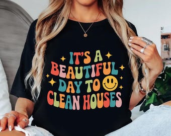 May include: Black t-shirt with the text "IT'S A BEAUTIFUL DAY TO CLEAN HOUSES" in colorful, retro-style lettering. The design includes a smiling face and star accents, perfect for house cleaners.