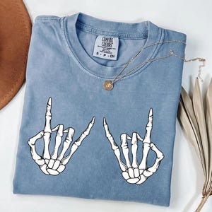 a blue t-shirt with a graphic design of two skeletal hands making a hand gesture, along with a necklace and a hat.