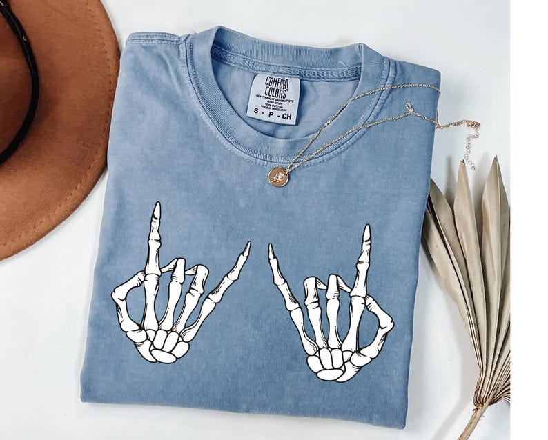 a blue t-shirt with a graphic design of two skeletal hands making a hand gesture, along with a necklace and a hat.