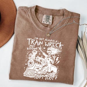 May include: A brown short-sleeve t-shirt featuring a white graphic print of a skeleton driving a train. The text reads: I'M NOT ALWAYS A TRAIN WRECK JUST KIDDING TOOT TOOT. The shirt has a crew neckline and a Comfort Colors brand label inside.