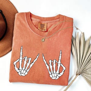 a red t-shirt with a graphic of two skeletal hands making a "rock on" sign, along with a brown hat and a fan.