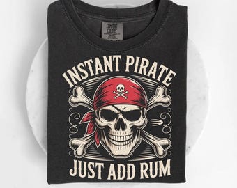 May include: Black t-shirt with a pirate graphic. The design includes a skull wearing a red bandana and crossed bones, with the text "INSTANT PIRATE JUST ADD RUM" in white. The shirt is folded.