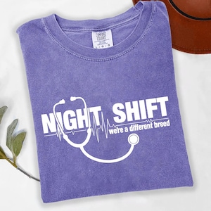 May include: A lavender Comfort Colors t-shirt with the words "NIGHT SHIFT" and "we're a different breed" in white, along with a stethoscope graphic. The shirt is folded and has a tag in the upper left corner.