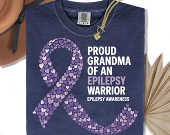 May include: Navy blue t-shirt with the text "PROUD GRANDMA OF AN EPILEPSY WARRIOR" and a purple ribbon made of hearts. The shirt also includes the words "EPILEPSY AWARENESS". A gold necklace with a heart pendant is draped on the shirt.