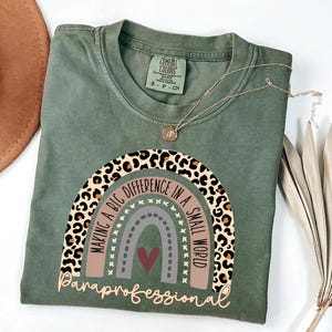Peut inclure: T-shirt vert olive avec un motif arc-en-ciel et le texte "MAKING A BIG DIFFERENCE IN A SMALL WORLD Paraprofessional". Le motif arc-en-ciel présente un imprimé léopard, marron, gris et un cœur rouge.