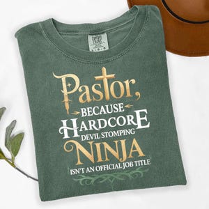 May include: A sage green t-shirt with the words "Pastor, because Hardcore Devil Stomping Ninja Isn't An Official Job Title" in gold and white lettering. The shirt has a crew neck and a Comfort Colors tag.