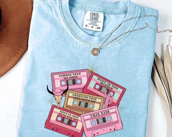 May include: A light blue t-shirt featuring a graphic print of four cassette tapes with the text "COWBOY TAKE ME AWAY," "MEET IN THE MIDDLE," "CARRYING YOUR LOVE WITH ME," "SHE'S IN LOVE WITH THE BOY," and "THIS KISS," plus a cow skull and cactus.