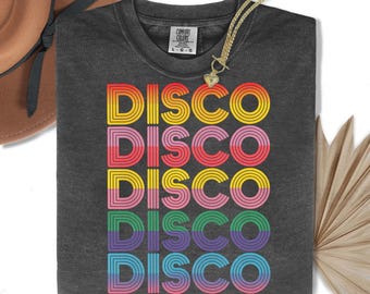 May include: Charcoal grey t-shirt featuring the word "DISCO" in a rainbow gradient, repeated five times. A gold necklace with a heart pendant rests on the shirt. A brown hat and dried plant are in the background.