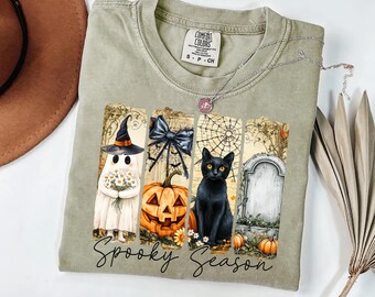 May include: A sage green t-shirt featuring a graphic print of a ghost in a witch hat holding flowers, a pumpkin, a black cat, and a tombstone. The text Spooky Season is printed in black script below the graphics. The shirt has a Comfort Colors label.
