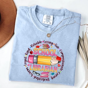 May include: A light blue t-shirt featuring a graphic of a yellow pencil, books, and glasses. The text reads "SCHOOL LIBRARIAN" surrounded by words like "Reliable," "Loving," "Fun," "Compassionate," "Caring," "Warm," "Loyal," and "Kind" in a circular design.