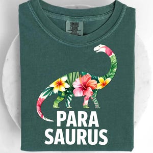 May include: Dark green t-shirt with a floral dinosaur design and the text "PARA SAURUS" in white. The dinosaur has a long neck and is decorated with tropical flowers and leaves. The shirt is folded.