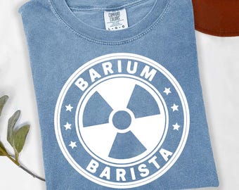 May include: A pale blue t-shirt featuring a white graphic with the words "BARIUM BARISTA" in a circular design, with a radiation symbol at the centre. The shirt has a crew neck and is made of a soft material.