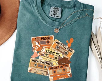 May include: A teal short-sleeve t-shirt featuring a graphic print of four cassette tapes labelled You Proof, Whiskey Glasses, Chasin' You, Talkin' Tennessee, and Wasted On You, decorated with illustrations of cowboy boots, a hat, and a cactus.