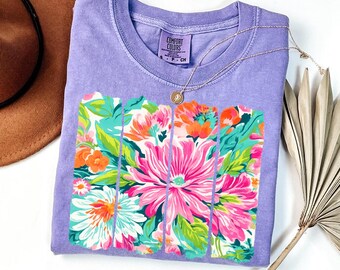 May include: A lavender short-sleeved t-shirt featuring a graphic print of four vertical panels filled with vibrant pink, orange, and teal floral patterns. The inner neck label displays the text Comfort Colors, size S, and 100% cotton.