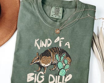May include: An olive green short-sleeve t-shirt featuring a graphic of an armadillo and a green cactus. The text reads "KIND OF A BIG DILLO" in cream-coloured lettering. The shirt has a Comfort Colors brand label at the inner neckline.