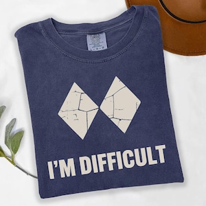 May include: A navy blue t-shirt with the text "I'M DIFFICULT" in white. The shirt has a graphic of two off-white diamond shapes. A brown hat is visible in the background.
