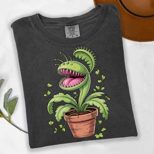 Puede incluir: Camiseta gris oscuro con un diseño de planta carnívora Venus en maceta de terracota. La planta tiene una cabeza verde con dientes afilados y una lengua rosa. La camiseta está doblada y tiene una etiqueta Comfort Colors.