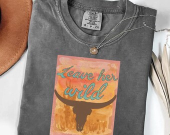 May include: A charcoal grey crew neck t-shirt featuring a rectangular graphic print with a watercolour-style orange and yellow background. The design includes a dark brown longhorn skull silhouette and the teal text Leave her wild.