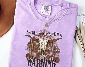 May include: A light purple short-sleeve t-shirt with a graphic print featuring a longhorn skull, desert cacti, and the text SHOULD'VE COME WITH A WARNING. The shirt has a Comfort Colors brand label on the inside collar showing size S.